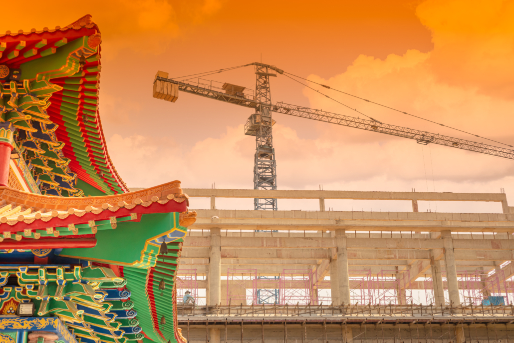 traditional Chinese building against new construction modernization of China city metro concept. A traditional Chinese temple roof with ornate, colourful detailing stands in contrast to a modern construction site featuring a crane and an unfinished concrete building, under an orange-hued sky.