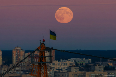 The Ukrainian flag atop a crane with a full moon rising behind city buildings at dusk.