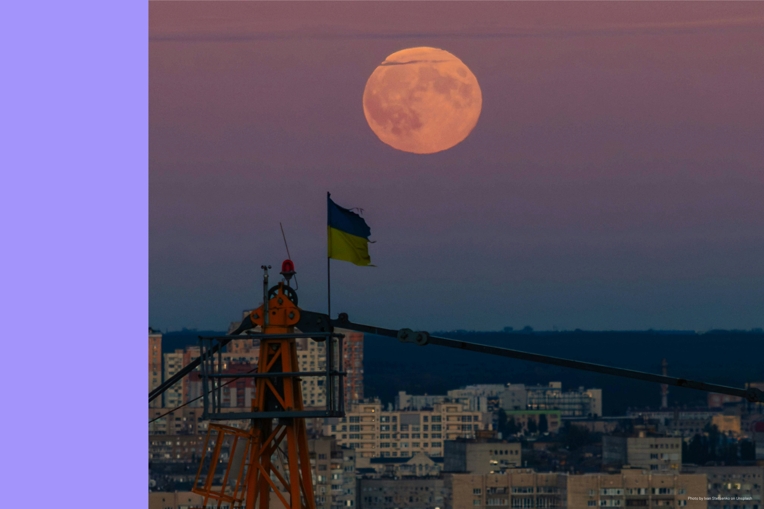 Ukrainian flag flying atop a crane against a city skyline at dusk with a large full moon rising in the background.